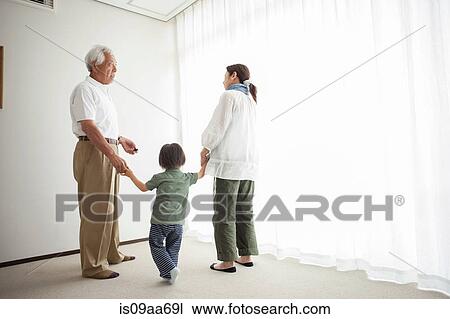 Stock Image - Three generation family standing by window holding hands. Fotosearch