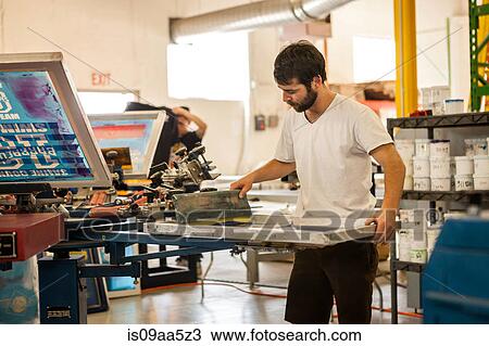 Stock Image - Worker pressing ink on frame in screen printing workshop. Fotosearch