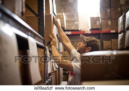 Stock Image - Worker reaching up for cardboard box stored in warehouse. Fotosearch