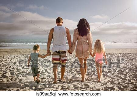 Stock Image - Young family holding hands together on beach, rear view. Fotosearch