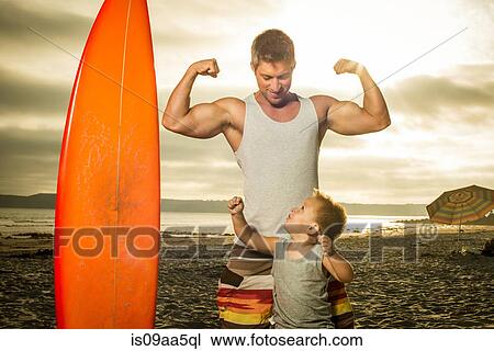 Stock Image - Young man and son flexing muscles on beach. Fotosearch