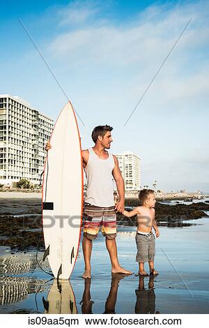 Picture - Young man holding surfboard with son in sea. Fotosearch