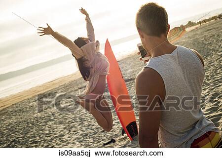 Stock Image - Young man photographing woman jumping on beach. Fotosearch