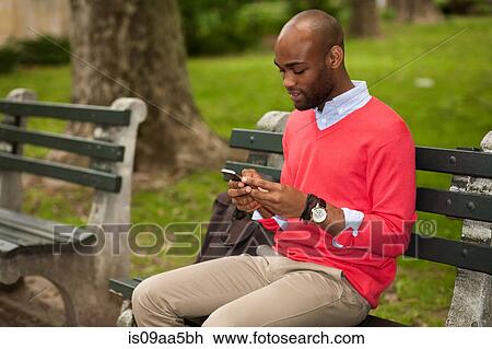 Stock Image - Young man sitting on park bench using mobile phone. Fotosearch