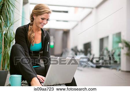 Stock Image - Young woman in gym corridor using laptop. Fotosearch