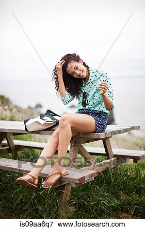Stock Photo - Young woman sitting on picnic table smiling, portrait. Fotosearch