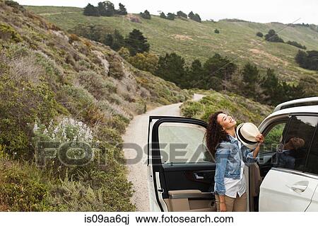 Stock Image - Young woman standing by car on country road, smiling. Fotosearch