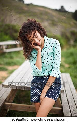 Stock Photography - Young woman with hand in hair standing by picnic table, smiling. Fotosearch