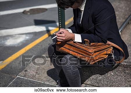 Stock Photo - Businessman sitting on kerb with satchel, smoking cigarette. Fotosearch