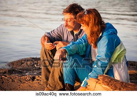 Stock Photography - Mature couple sitting lakeside with mobile phones, Bath, Maine, USA. Fotosearch