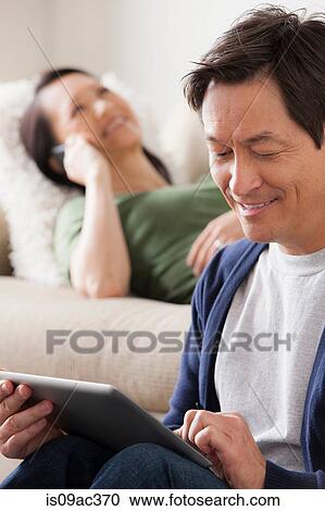 Stock Image - Mature man using digital tablet, woman lying on sofa. Fotosearch