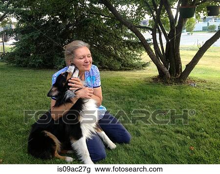 Stock Image - Mature woman kneeling on grass with dog, smiling. Fotosearch
