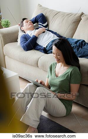 Stock Photograph - Mature woman using digital tablet, man asleep on sofa. Fotosearch