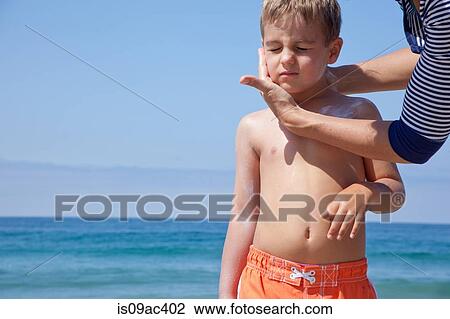 Stock Image - Mother applying sun cream to sons face at beach. Fotosearch