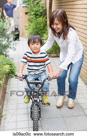 Stock Image - Mother helping son to ride a bike. Fotosearch