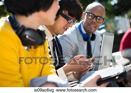 Stock Image - Three casual businesspeople using laptop in park. Fotosearch