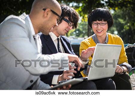 Stock Image - Three casual businesspeople using laptop in park. Fotosearch