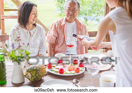 Stock Image - Woman serving wine to parents at dining table. Fotosearch