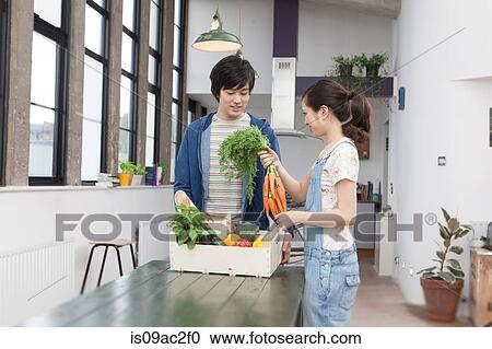 Stock Image - Young couple in kitchen with box of vegetables holding carrots. Fotosearch