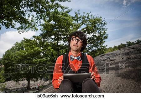 Stock Image - Young man sitting on rocks using tablet. Fotosearch