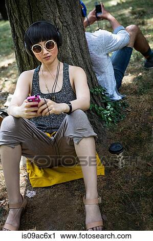 Stock Image - Young woman leaning against tree trunk with mp3 player. Fotosearch