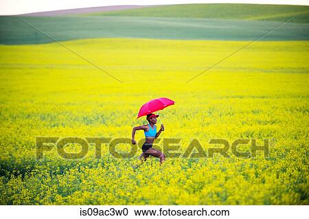 Stock Image - Young woman running in oil seed rape field with red umbrella. Fotosearch