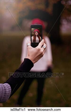 Stock Image - Hand holding smartphone, taking photograph of young woman. Fotosearch