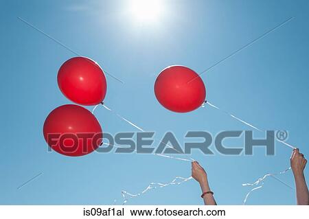 Stock Image - Hands holding three red balloons against blue sky. Fotosearch