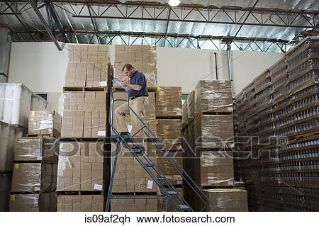 Stock Image - Man on ladders in warehouse with cardboard boxes. Fotosearch
