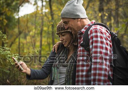 Stock Image - Mature couple taking self portrait photograph on smartphone in forest. Fotosearch