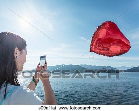 Stock Image - Mature woman photographing heart shaped balloon. Fotosearch