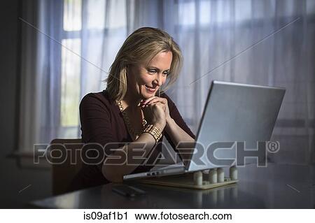 Stock Image - Mature woman sitting at home using laptop. Fotosearch
