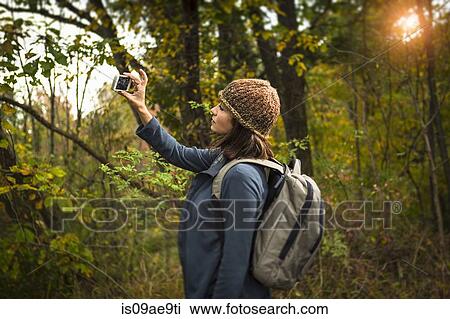 Stock Image - Mature woman taking photograph in forest using smartphone. Fotosearch