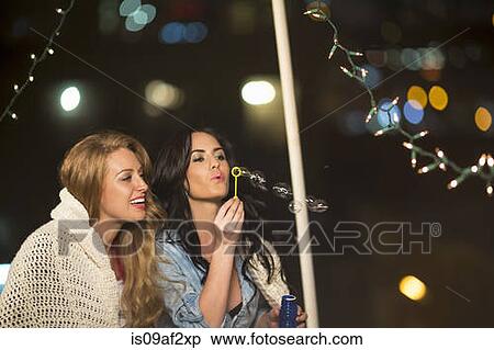 Stock Photography - Two female friends blowing bubbles at rooftop party. Fotosearch