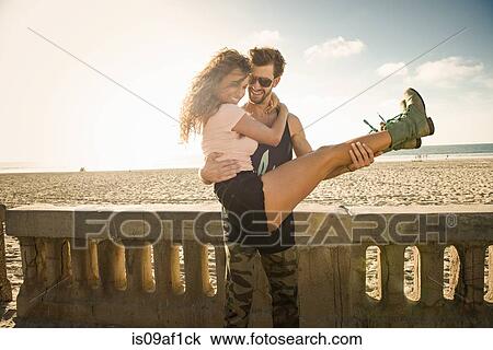 Stock Image - Young man holding girlfriend in arms at San Diego beach. Fotosearch