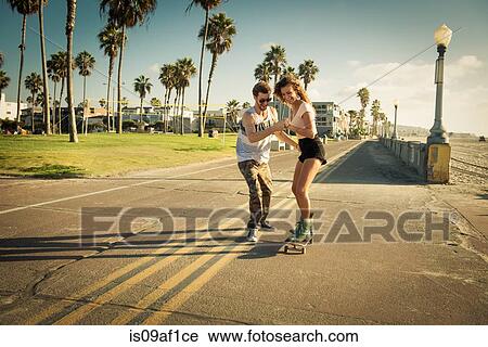 Stock Image - Young woman on skateboard at San Diego beach, boyfriend helping. Fotosearch