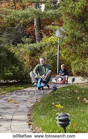 Stock Image - Father and young son riding on toy cars in garden. Fotosearch