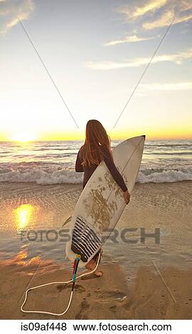 Stock Photo - Girl at beach carrying surfboard wearing wetsuit. Fotosearch
