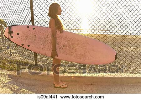 Stock Image - Girl standing at beach holding pink surfboard. Fotosearch