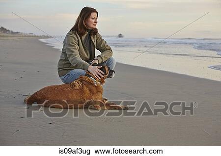 Stock Image - Mature woman petting her dog on beach. Fotosearch