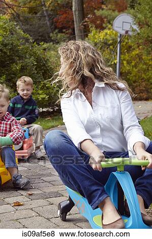 Stock Photo - Mother and young sons riding on toy cars in garden. Fotosearch
