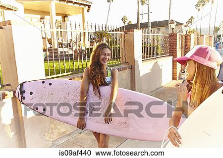 Stock Image - Two girls carrying surfboards laughing and smiling. Fotosearch