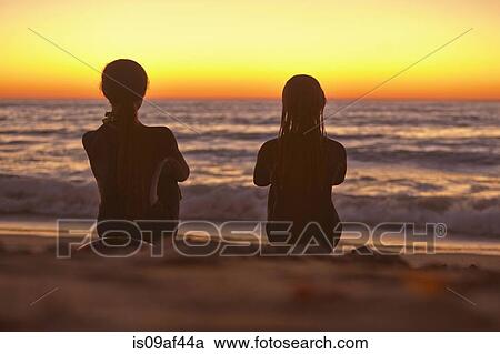 Stock Image - Two girls sitting on beach at sunset looking out to sea. Fotosearch