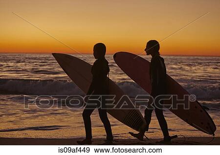 Two girls walking along beach at sunset carrying surfboards View Large Photo Image Stock Photo - Two girls walking along beach at sunset carrying surfboards. Fotosearch