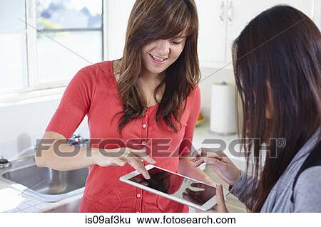 Stock Photograph - Two young women in kitchen playing game on digital tablet. Fotosearch