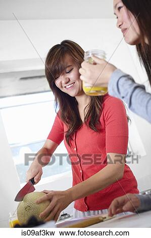 Stock Photo - Two young women in kitchen slicing melon. Fotosearch