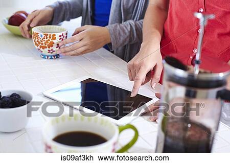 Stock Image - Two young women in kitchen using digital tablet whilst having coffee. Fotosearch