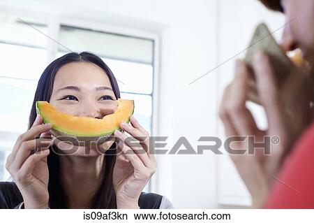 Picture - Two young women in kitchen with melon mouths. Fotosearch
