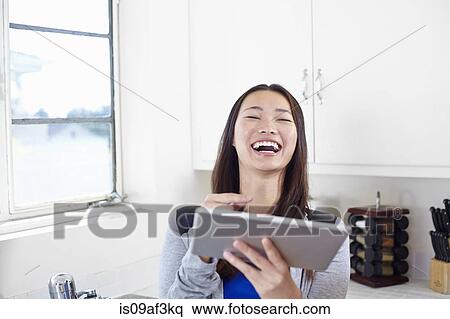 Stock Photography - Young woman in kitchen laughing at digital tablet. Fotosearch