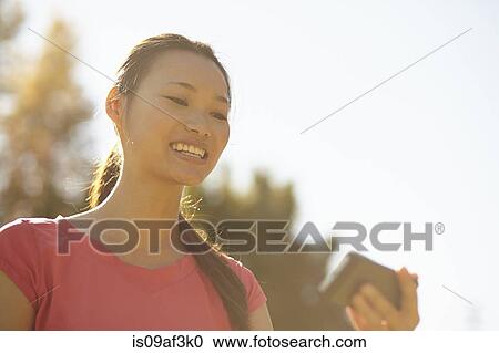 Stock Image - Young woman in park looking at mobile phone. Fotosearch